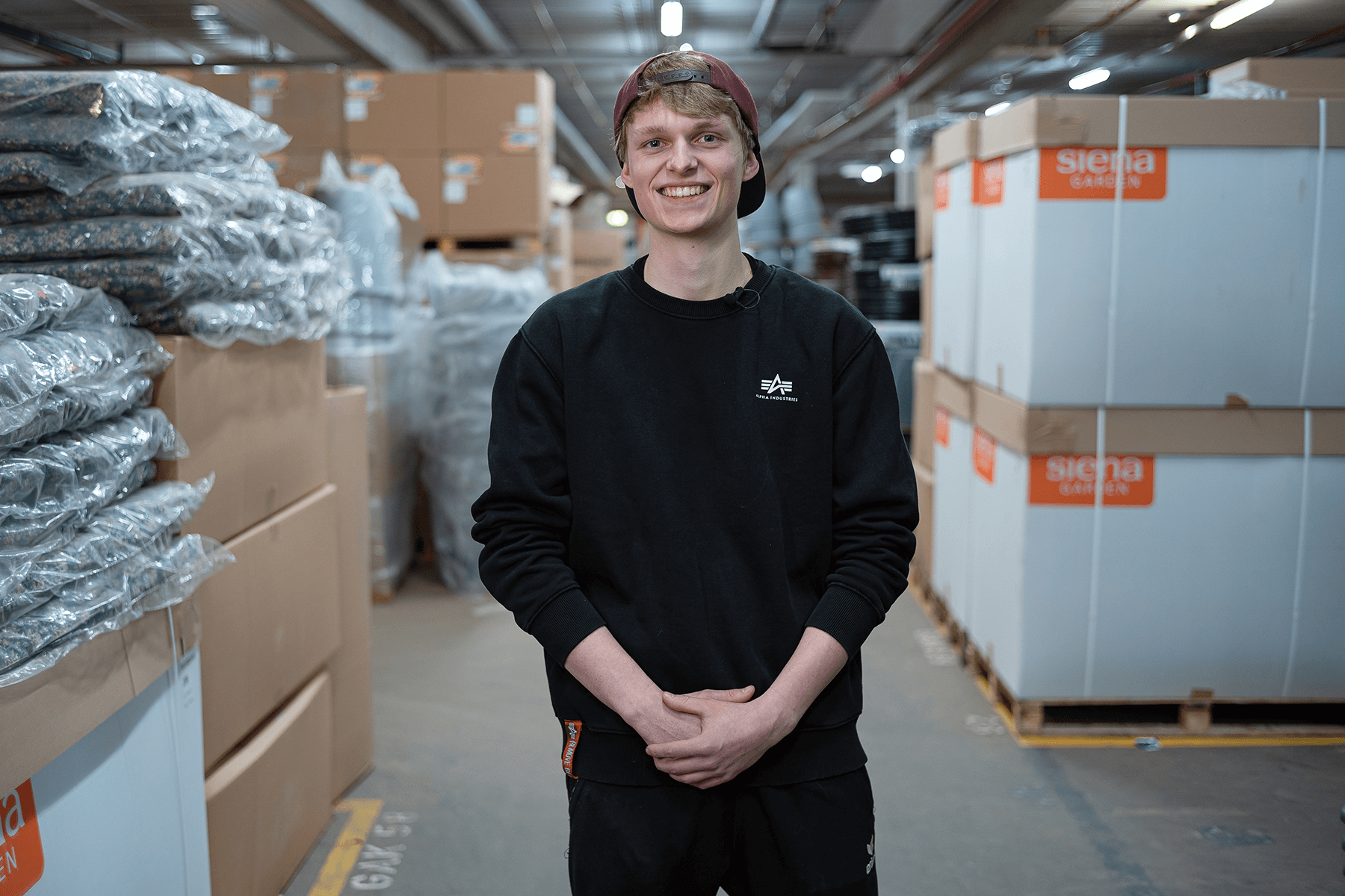 Mitarbeiter im Lager von SIENA GARDEN, steht lächelnd zwischen Paletten mit verpackter Ware.